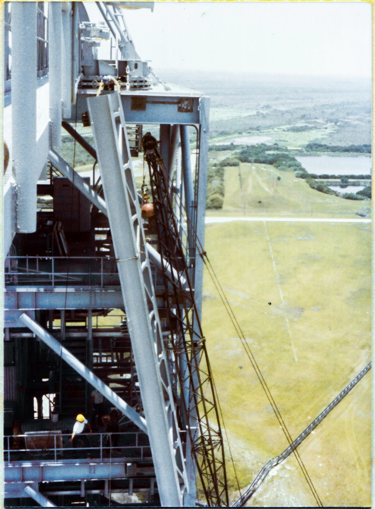 Image 082. Leaning dangerously out over empty space from a kneeling position at Elevation 300'-0”, unharnessed, untethered, Union Ironworker James Dixon barehands the top of the GOX Arm Hinges Support Strongback where it meets the top of the Fixed Service Structure at Space Shuttle Launch Complex 39-B, Kennedy Space Center, Florida, as another, unknown alas, Union Ironworker in a white shirt wearing a yellow hardhat, two levels below at Elevation 260'-0”, gives hand signals to the crane operator, at the controls in his cab, down on the Pad Deck at Elevation 53'-0”. The crane operator has no way to see what's going on far above him with any kind of detail, and relies exclusively on the hand signals he's being given, even as everyone else relies on his skill at swinging, rotating, and nudging forty feet of steel truss weighing well over 10,000 pounds into a final position that has to wind up within roughly one sixteenth of an inch of where it's needed for final attachment to the FSS. Everyone's lives hang in the balance as this work is done, they all know this, and they all trust one another. With their own lives. As they do the work. Photo by James MacLaren.
